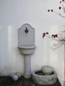 Ein Steinbrunnen, der an einer Wand befestigt ist, mit einem Auffangbecken für Wasser und darüber ein Wasserhahn als Wasserquelle.